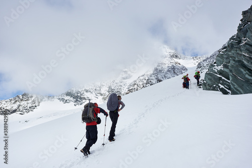 Male travelers with backpacks walking uphill in winter mountains. Group of alpinists with trekking sticks walking through snow and heading to mountaintop. Concept of hiking and mountaineering.