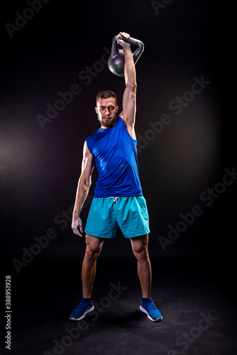 A sports man in a blue T-shirt and shorts trains with a kettlebell on a black background.