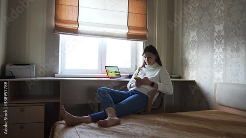 Sits on chair, home in room, white sweater blue jeans. Laptop table. Fills in form on phone, payment for services, calendar entries, work correspondence in chat. Looks clock, waiting for the meeting.