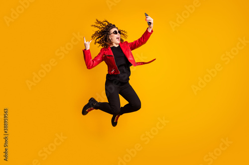 Fototapeta Naklejka Na Ścianę i Meble -  Full length body size view of cheerful wavy-haired girl jumping using cell showing horn sign taking selfie isolated on yellow color background