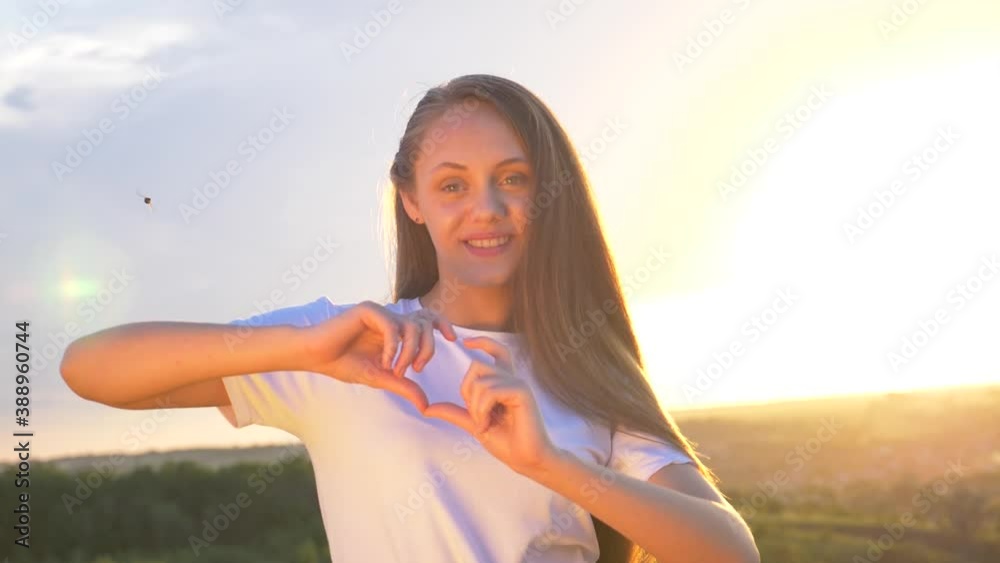 A girl at sunset looks into the camera and makes a heart symbol out of her fingers. Heart shape with hands, a sign of love. Concept of health care, vacation, help, Valentine's day