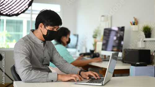 A young man photographer with face mask working with mock up laptop in comfortable workspace.