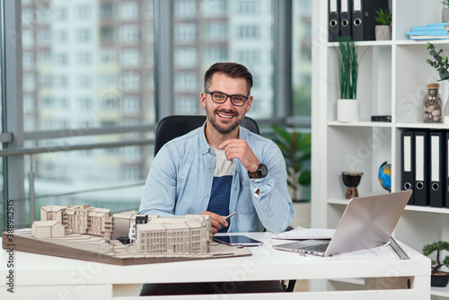 Likable smiling successful bearded 35-aged architect in glasses sits at his workplace with mock-up of future buildings. photo