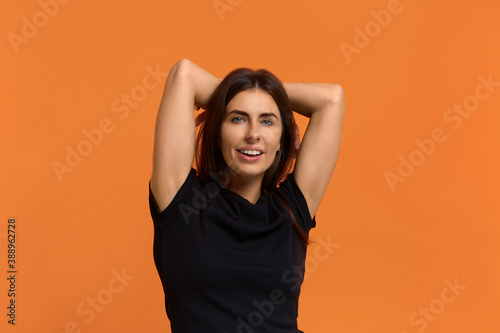 Relax with me. Relaxed carefree caucasian woman in black t-shirt keeps hands behind head, has positive expression, smiles broadly looking at camera. Isolated over an orange background. © 5M
