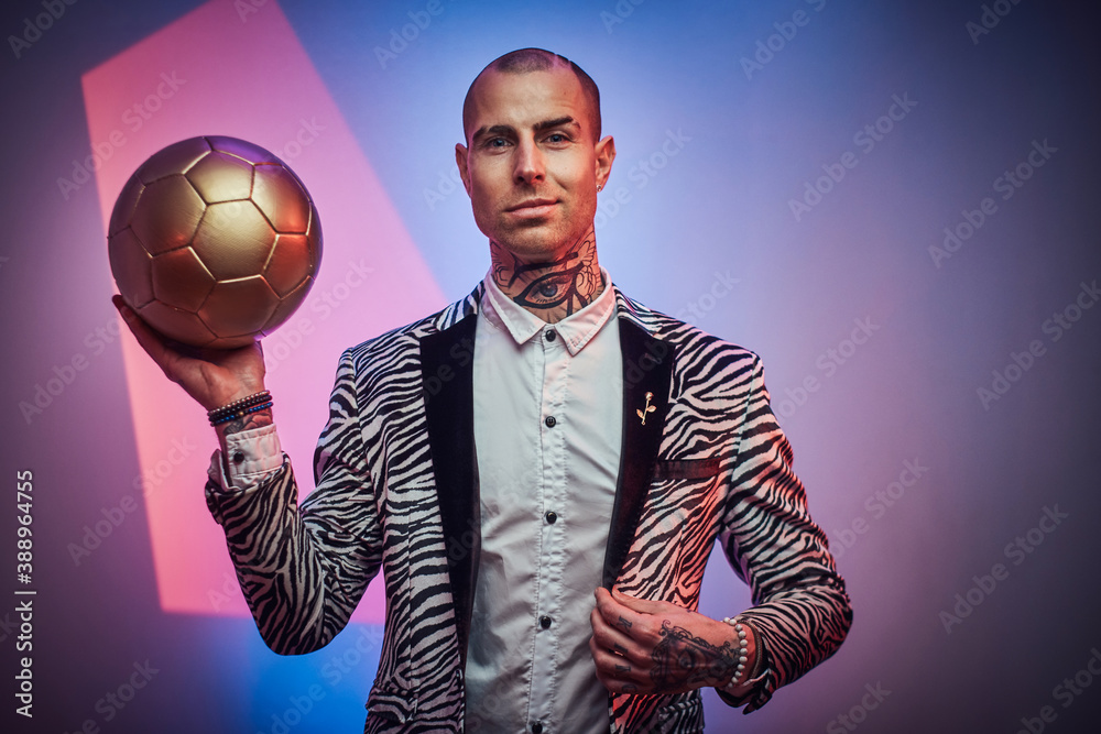 Plakat Positive and succesful football trainer dressed in custom suit with jewellery poses with golden ball in abstract light background.