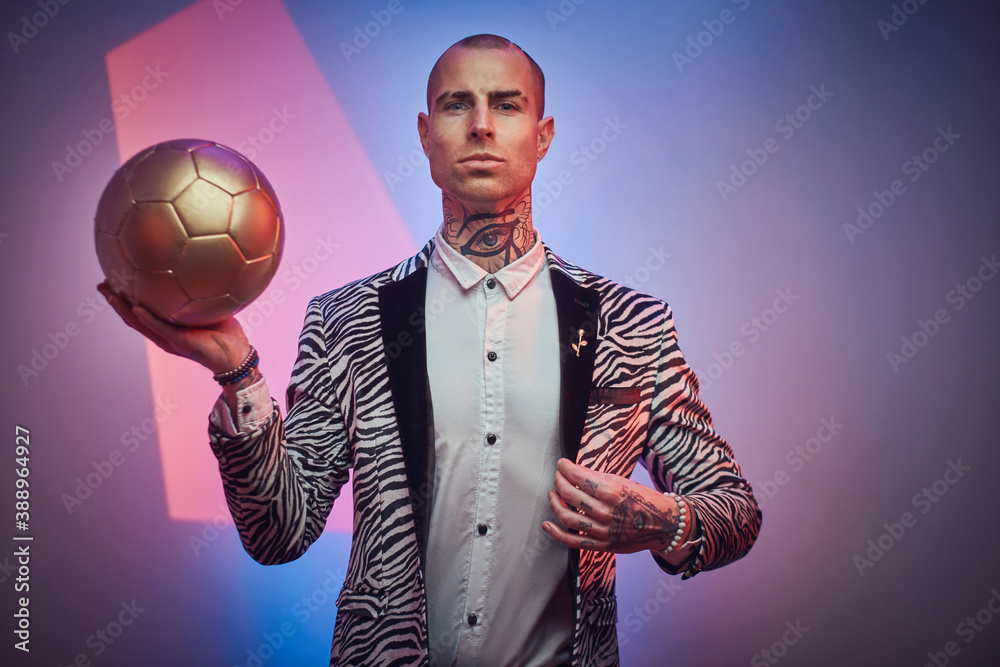 Plakat Serious tattooed guy with jewellery in stylish suit poses in colored abstract background holding golden ball.
