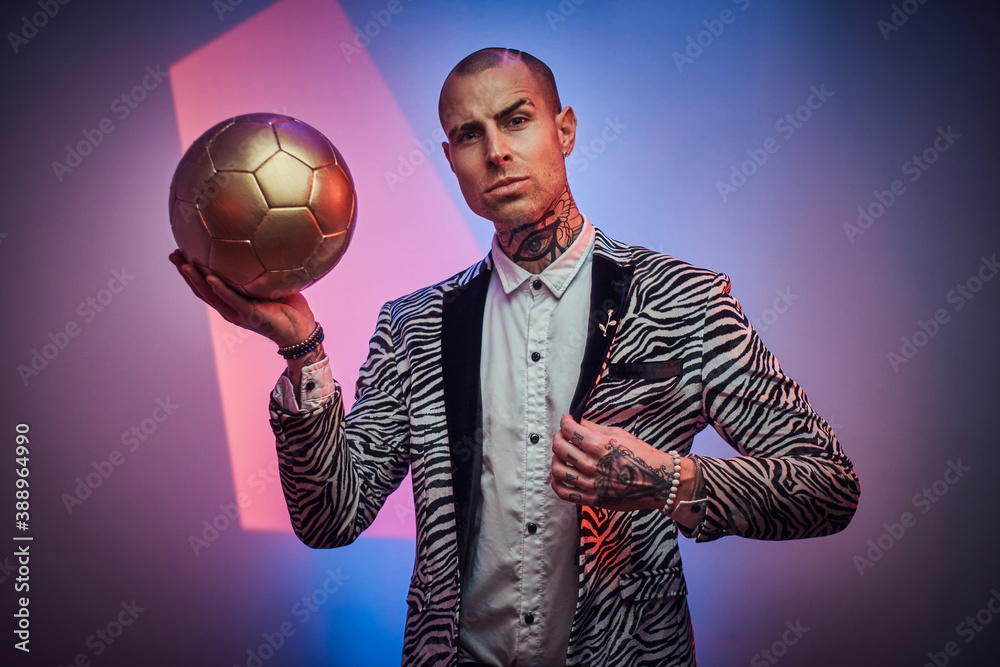 Plakat Stylish and succesful football trainer dressed in custom suit with jewellery poses with golden ball in abstract light background.