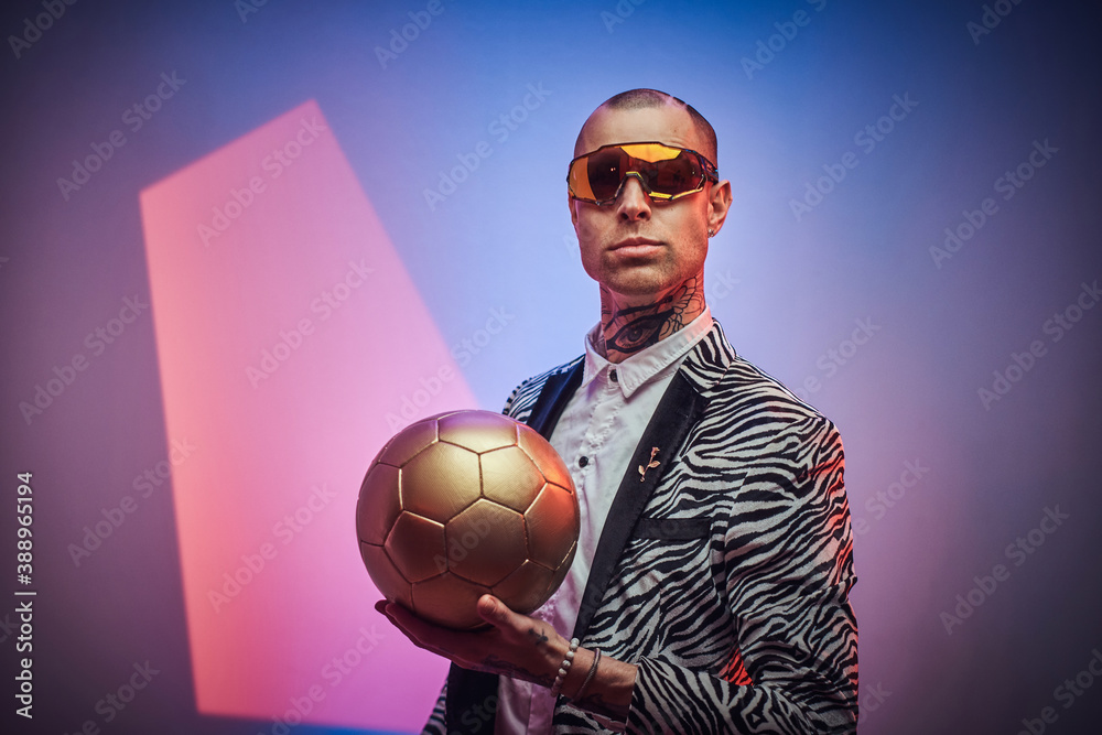 Plakat Handsome guy dressed in custom suit with short haircut and eyewears poses with golden soccer ball in abstract background.