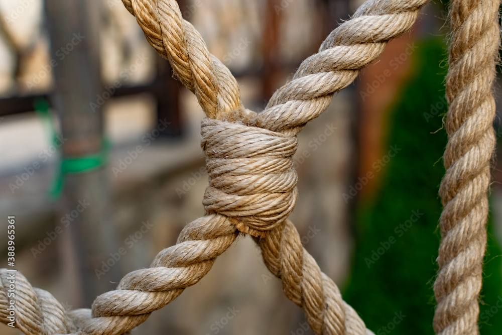 Thick rope ropes, intertwined crosswise, hang on wooden railings, decorative fence along the alley in the Park for recreation. Metal rings and large strong knots on a stone background