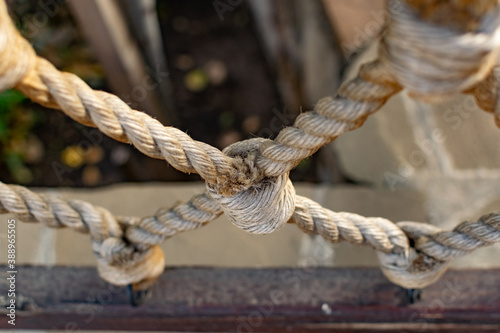 Thick rope ropes, intertwined crosswise, hang on wooden railings, decorative fence along the alley in the Park for recreation. Metal rings and large strong knots on a stone background