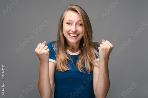 Successful Young female is smiling happily, making Winner gesture, standing gray background