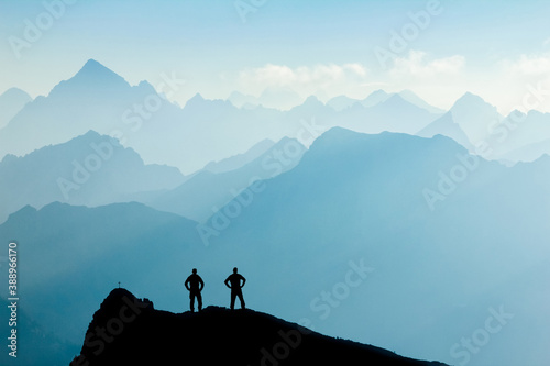 Two Men reaching summit after climbing and hiking enjoying freedom and looking towards mountains silhouettes panorama during sunrise.