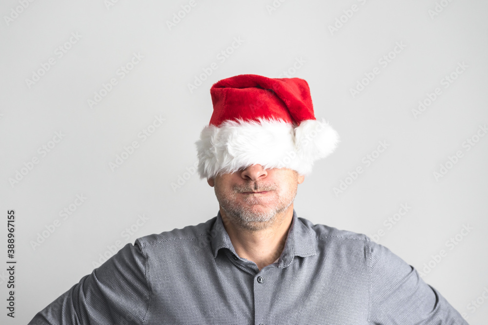 Gestresster Mann mit Weihnachtsmütze über den Augen vor einem neutralen Hintergrund. Graues Hemd, Portrait, über 50. Gestresster Mann mit Weihnachtsmütze über den Augen vor einem neutralen Hintergrund. Graues Hemd, Portrait, über 50.