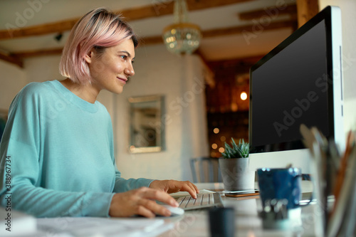 Attractive young woman copywriter typing on keyboard of desktop computer, sitting at her workplace. Skilled stylish female web designer enjoying creative occupation, working remotely from home