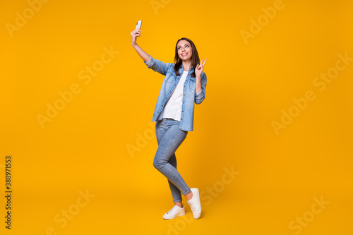 Full length photo of attractive funny lady good mood hold telephone making selfies youth blog show v-sign symbol wear casual denim shirt sneakers isolated vivid yellow color background