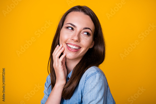 Closeup photo of attractive cheerful lady good mood beaming smile look interested side empty space touch arm cheek wear casual denim shirt isolated vivid yellow color background