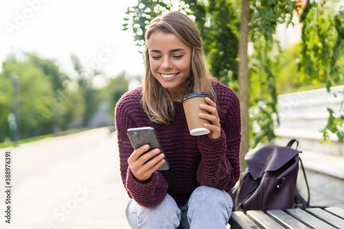 Beautiful smiling young s woman sitting on a bench outdoors, using mobile phone while drinking takeaway coffee cup