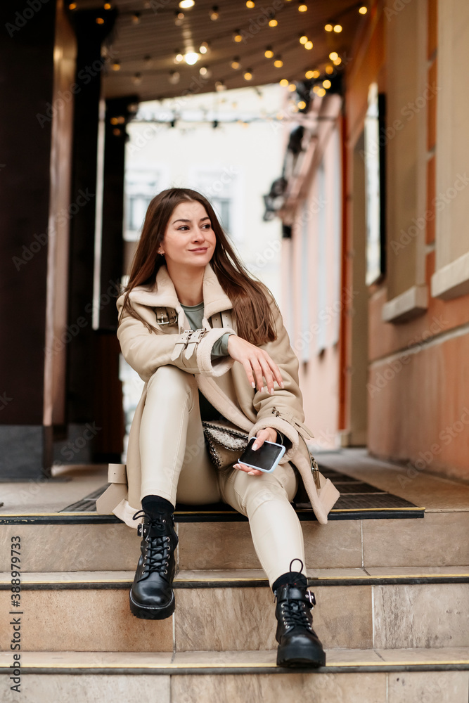 A young woman sits on the steps of a building in the city.Portrait of a beautiful woman with a phone in swirls against the background of a garland of light bulbs. Christmas and New Year concept. A young woman sits on the steps of a building in the city.Portrait of a beautiful woman with a phone in swirls against the background of a garland of light bulbs. Christmas and New Year concept.