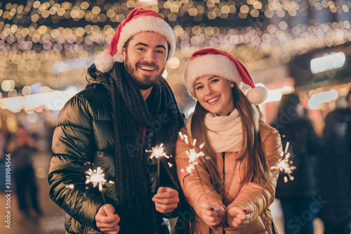 Photo of couple students celebrate christmas hold sparklers under x-mas evening outside illumination wear santa claus cap
