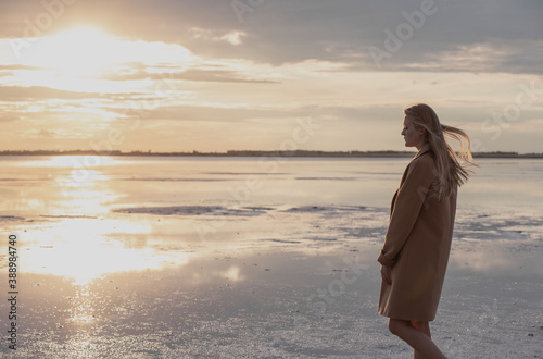 Beautiful young blonde girl with long hair walks along the lake shore and looks at the sunset. Autumn walk. The girl in the coat