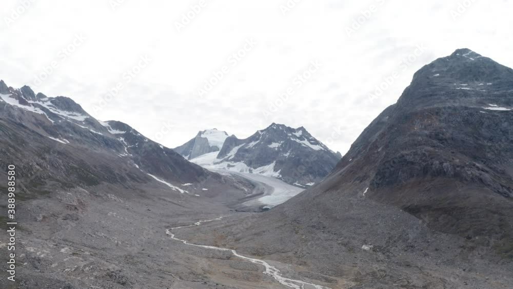 Glacier between mountain peaks in Greenland. Snow capped mountains and glacial melt river flowing downhill toward the sea. Bright sky above rugged terrain carved by ancient ice movement. Aerial view. Glacier between mountain peaks in Greenland. Snow capped mountains and glacial melt river flowing downhill toward the sea. Bright sky above rugged terrain carved by ancient ice movement. Aerial view.