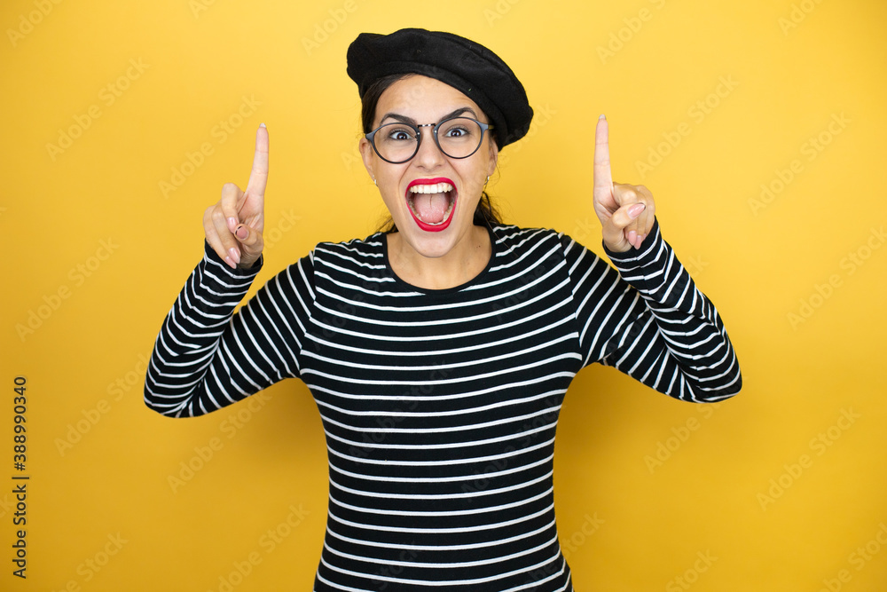 Young beautiful brunette woman wearing french beret and glasses over yellow background amazed and surprised looking at the camera and pointing up with fingers and raised arms