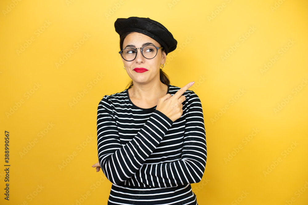 Young beautiful brunette woman wearing french beret and glasses over yellow background smiling happy pointing with hand and finger to the side