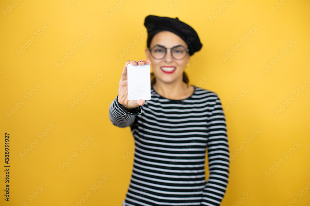 Young beautiful brunette woman wearing french beret and glasses over yellow background smiling and holding white card Young beautiful brunette woman wearing french beret and glasses over yellow background smiling and holding white card