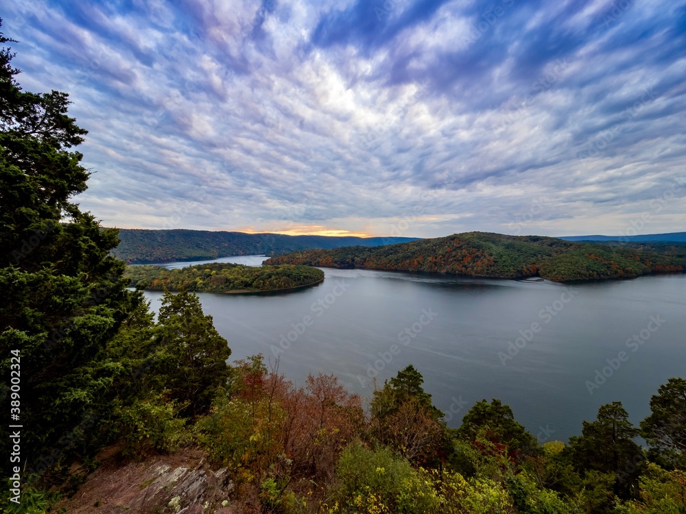 View of Raystown Lake in Pennsylvania in the Fall from Hawn’s Overlook.  The water is smooth as glass, colorful trees in the distance and a dramatic sunset sky filled with blue, white and pink.
