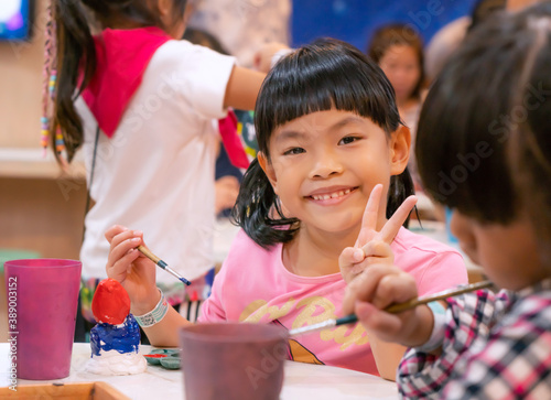 Portrait Asian child girl in art workshop, happy and smiling face, show gesture two fingers, cute face with black hair and bang. Blurred classmate foreground and background.