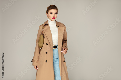 Fashionable young model posing against grey background dressed in trench coat and holding stylish  bag. Autumn fashion concept photo