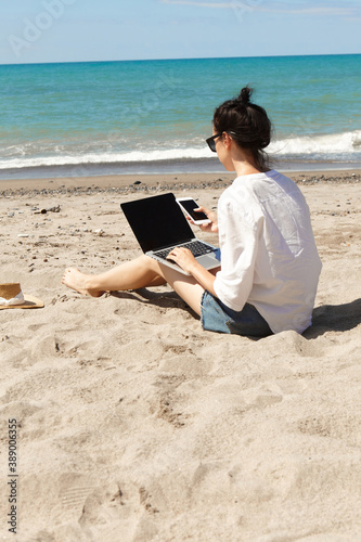 Young woman using laptop computer and holding a cell phone on a beach. Vacation lifestyle communication. Freelance work concept