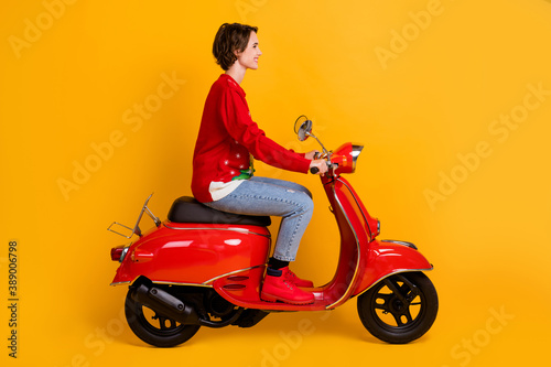 Full length profile side photo of positive girl ride red scooter travel x-mas time eve noel party wear christmas tree decor pullover denim jeans isolated bright shine color background