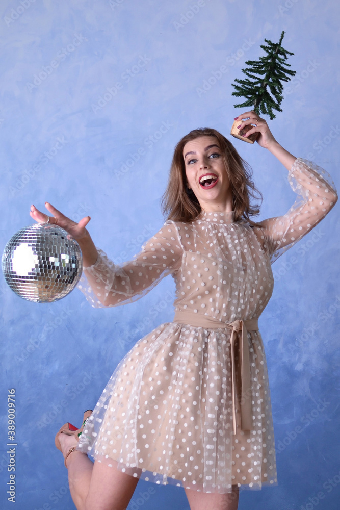 Naklejka premium beautiful happy woman in a beige dress holding a shiny disco ball and a small Christmas tree on a blue background. Holiday concept, new year, christmas, party.