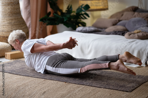 Senior woman practicing yoga at home indoor, doing Salabhasana or Locust pose. photo