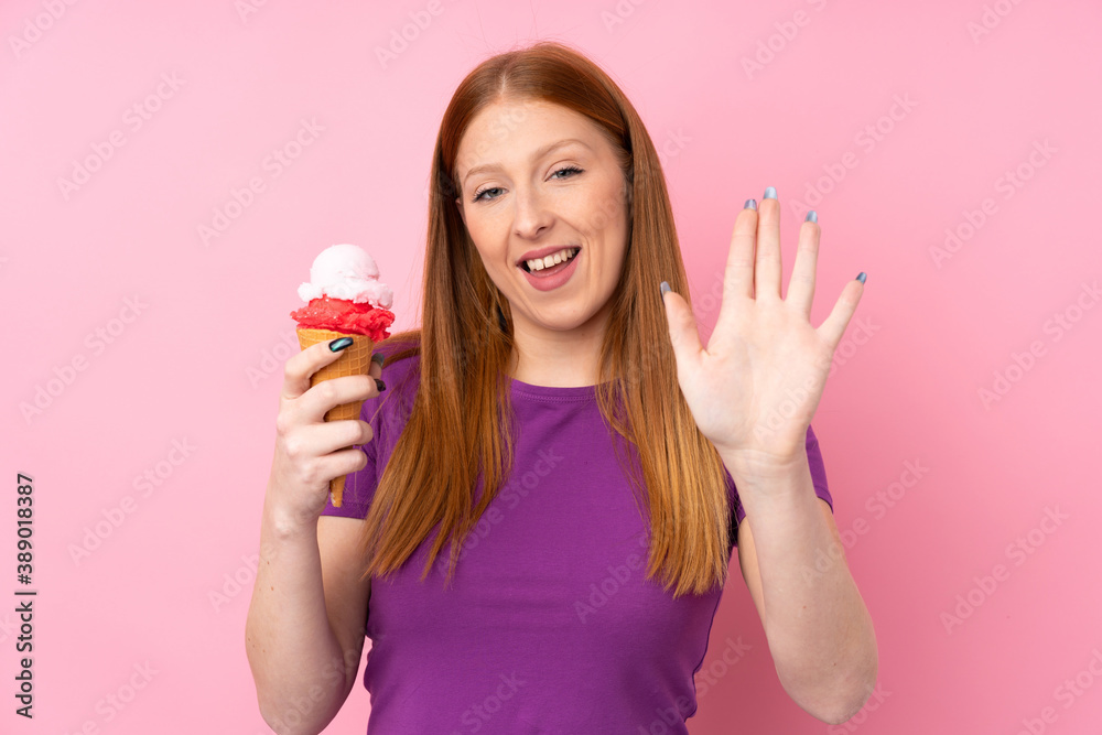Young redhead woman with a cornet ice cream over isolated pink background saluting with hand with happy expression Young redhead woman with a cornet ice cream over isolated pink background saluting with hand with happy expression