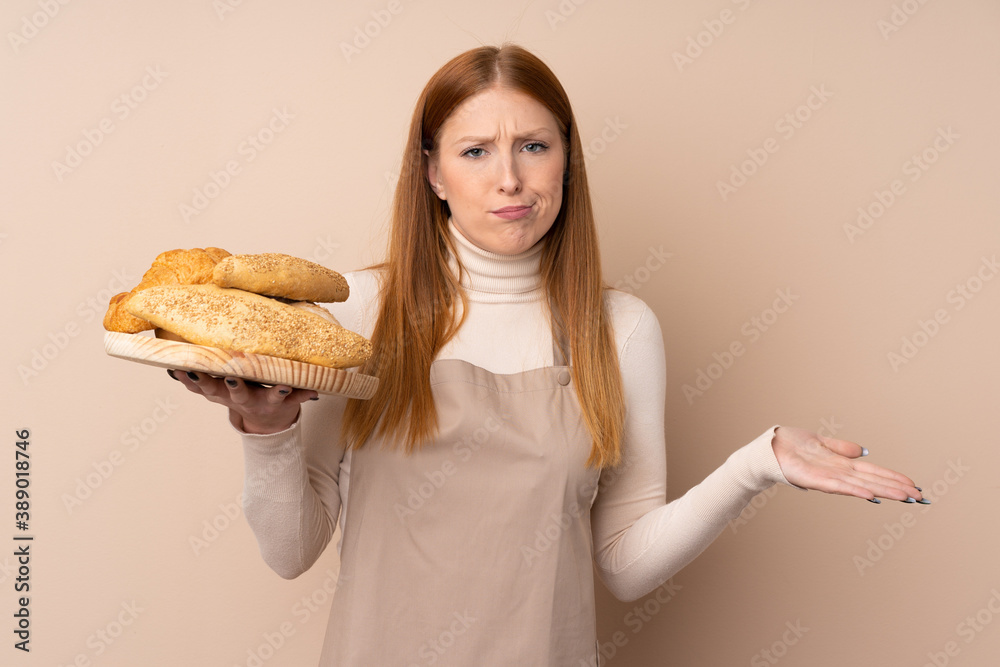 Young redhead woman in chef uniform. Female baker holding a table with several breads making doubts gesture while lifting the shoulders Young redhead woman in chef uniform. Female baker holding a table with several breads making doubts gesture while lifting the shoulders
