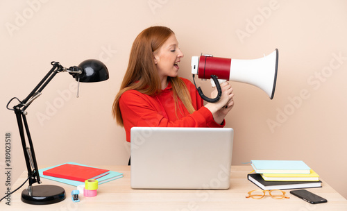 Young student woman in a workplace with a laptop shouting through a megaphone