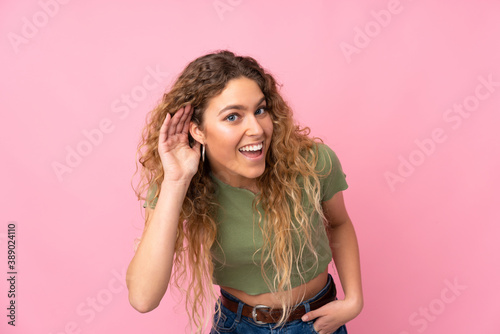 Young blonde woman with curly hair isolated on pink background listening to something by putting hand on the ear