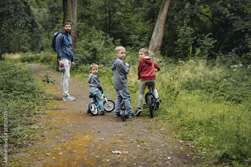 A young caucasian bearded man and three cute boys walking along a path in forest in summer time. One boy is cycling by bicycle, second by runbike, third is riding a kick scooter