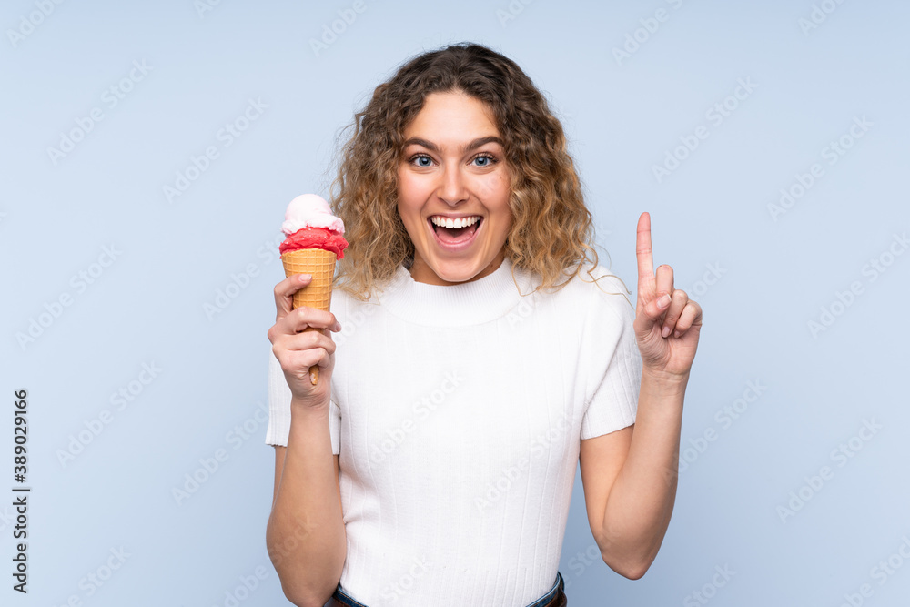 Young blonde woman with curly hair holding a cornet ice cream isolated on blue background pointing up a great idea