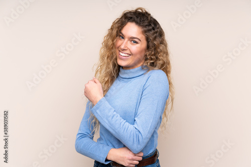 Young blonde woman with curly hair wearing a turtleneck sweater isolated on beige background celebrating a victory