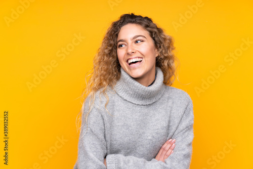 Young blonde woman with curly hair wearing a turtleneck sweater isolated on yellow background happy and smiling © luismolinero