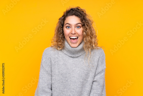 Young blonde woman with curly hair wearing a turtleneck sweater isolated on yellow background with surprise facial expression © luismolinero