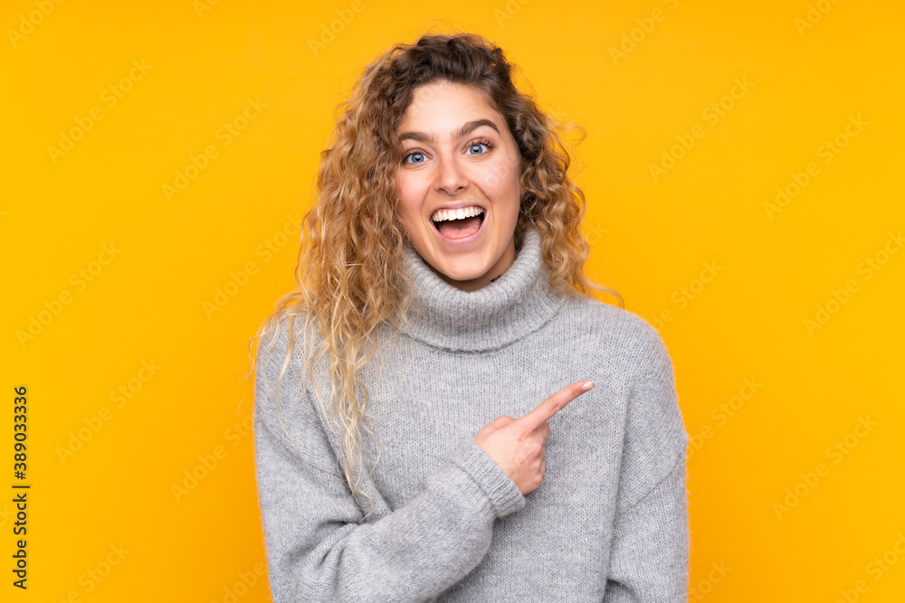Young blonde woman with curly hair wearing a turtleneck sweater isolated on yellow background surprised and pointing side