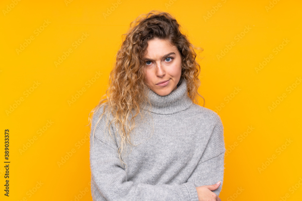 Young blonde woman with curly hair wearing a turtleneck sweater isolated on yellow background feeling upset