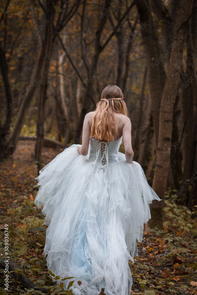 A girl-bride in a long white dress walks through the autumn forest, view from the back.