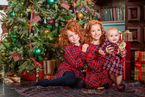 Red-haired curly children play near the Christmas tree next to gift boxes.