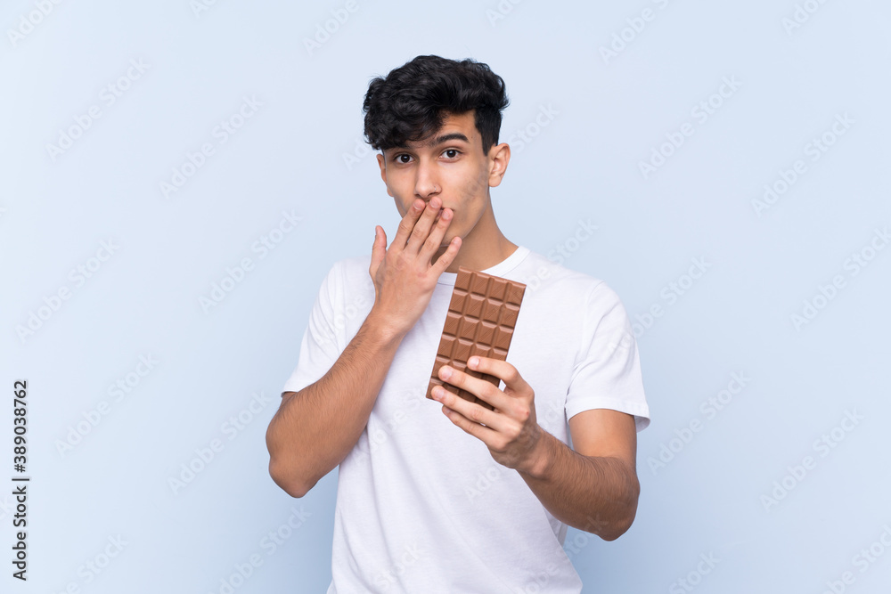 Young Argentinian man over isolated blue background taking a chocolate tablet and surprised Young Argentinian man over isolated blue background taking a chocolate tablet and surprised