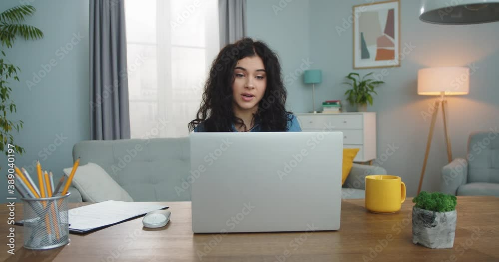 Beautiful young woman working on computer and showing clipboard to web camera in home office room. Attractive Caucasian female using laptop to talk with collegue in video conference indoors. Beautiful young woman working on computer and showing clipboard to web camera in home office room. Attractive Caucasian female using laptop to talk with collegue in video conference indoors.